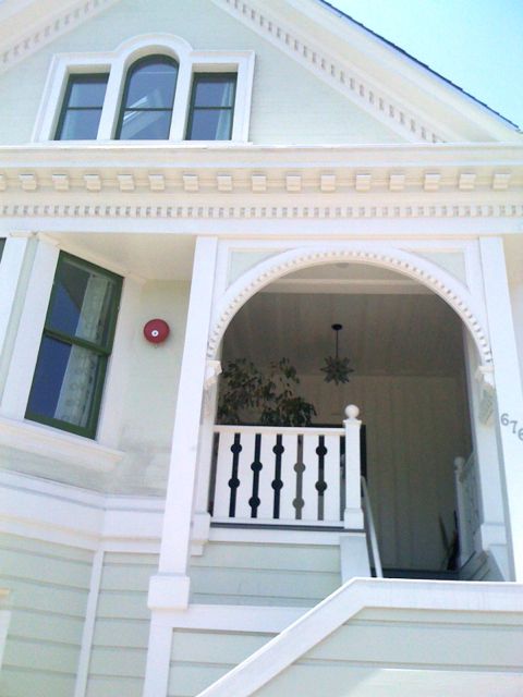 A light-colored, two-story house with a front porch and an arched entrance. The porch features a railing with balusters, a hanging light fixture, and a potted plant.